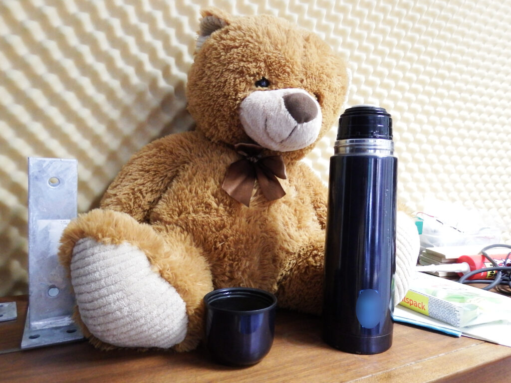 Ein Teddybär sitzt vor einer schallgedämpften Wand, davor steht eine offene Thermoskanne.