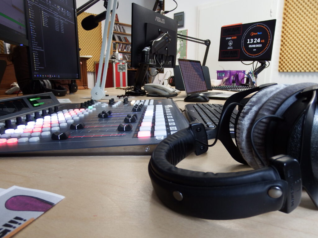 Foto: Nahaufnahme eines Studiopultes, im Vordergrund liegt ein paar Kopfhörer auf dem Tisch. Im Hintergrund: Monitore und Mikrofone.
