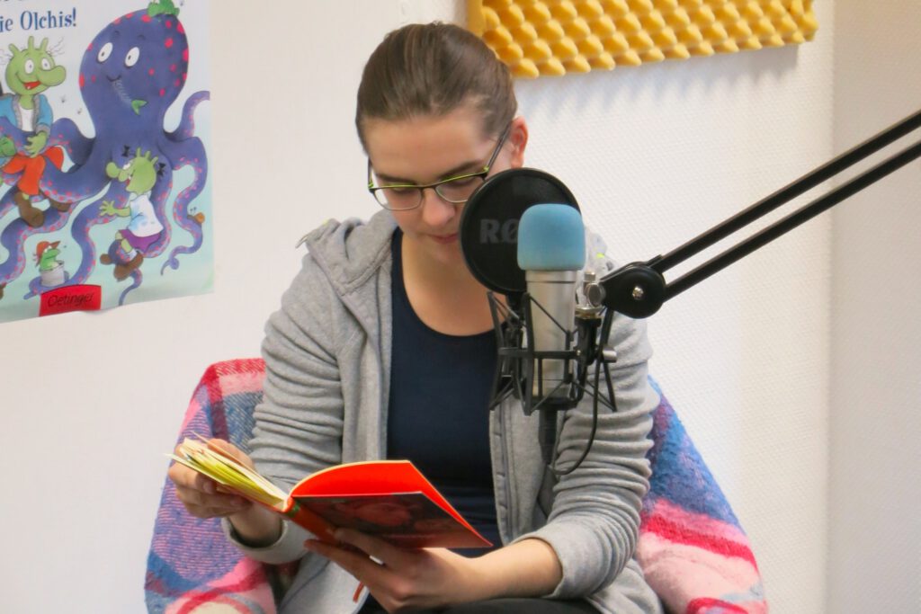 Weiblich gelesene Person sitzt hinter einem Mikrofon. Sie hält ein Buch in den Händen und liest daraus vor.
