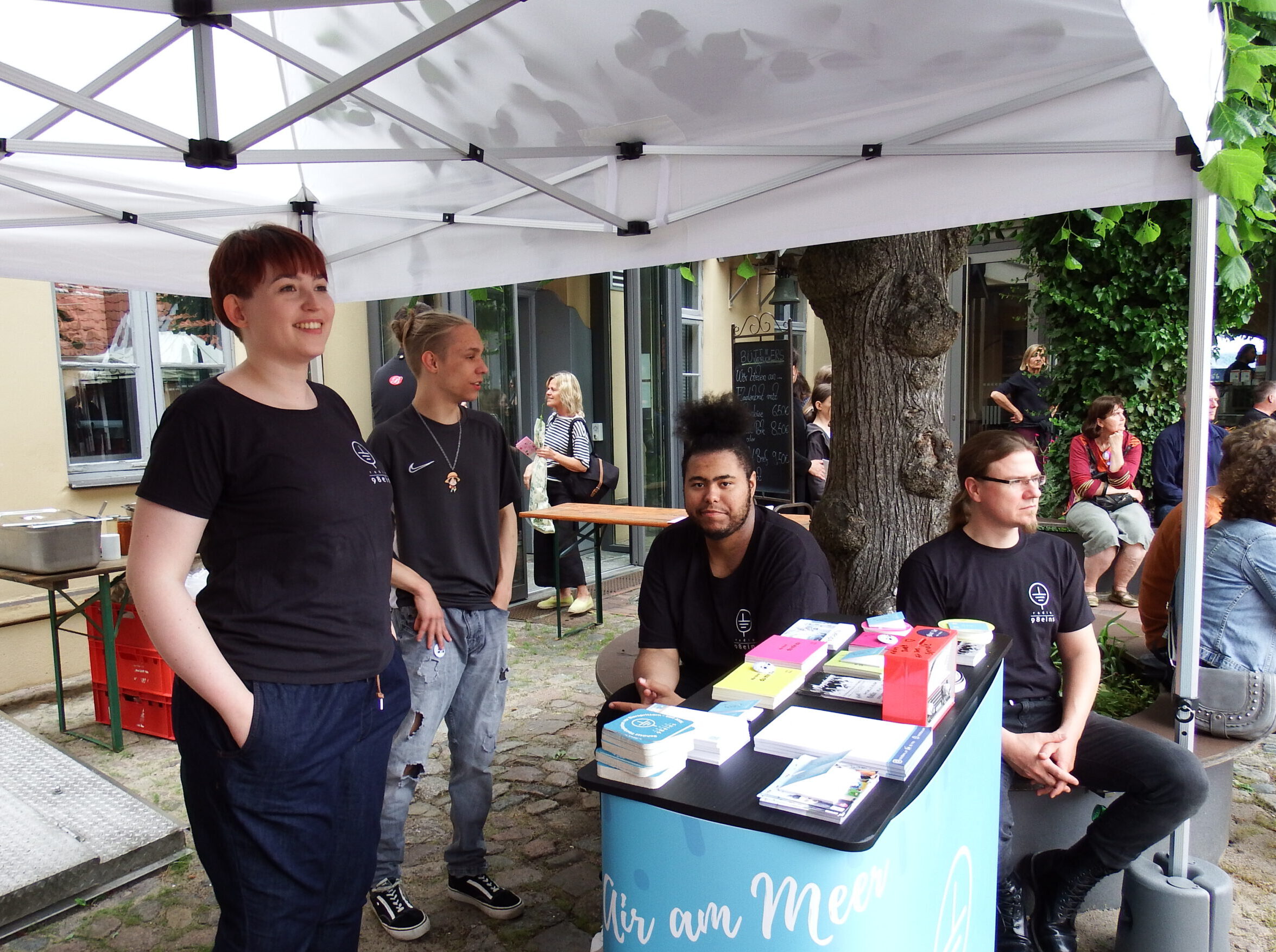 Der radio 98eins PR Stand im St. Spiritus Innenhof. An einer hellblauen Messetheke steht im Vordergrund eine Radionautin mit kurzen roten Haaren, in einem schwarzen radio 98eins Crew-Shirt. Auf der Messetheke liegen bunte Mechartikel, u.a. Postkarten, Bleistifte, Sticker, und Flyer. Im Hintergrund sitzen zwei weitere Radiomitglieder auf einer Bank. Ein jüngerer Praktikant steht daneben. Am oberen Bildrand sieht man den weißen Pavillon, der den PR Stand überdacht.