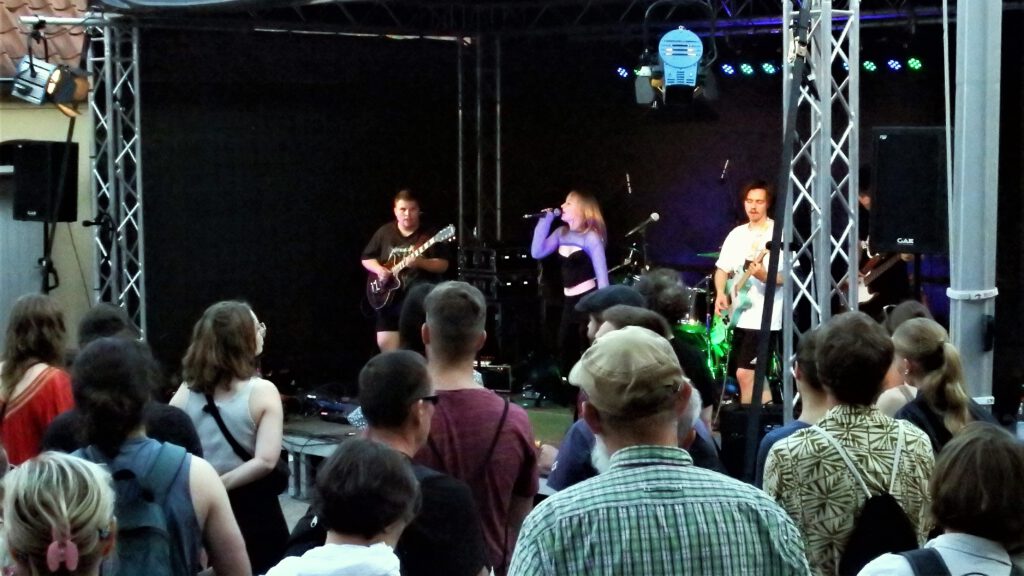 Foto einer Bühne aus dem Publikum heraus, im Vordergrund Zuschauende. Dahinter sind drei Bandmitglieder zu sehen, die äußeren spielen Bass oder E-Gitarre, die Sängerin in der Mitte springt leicht in die Luft.