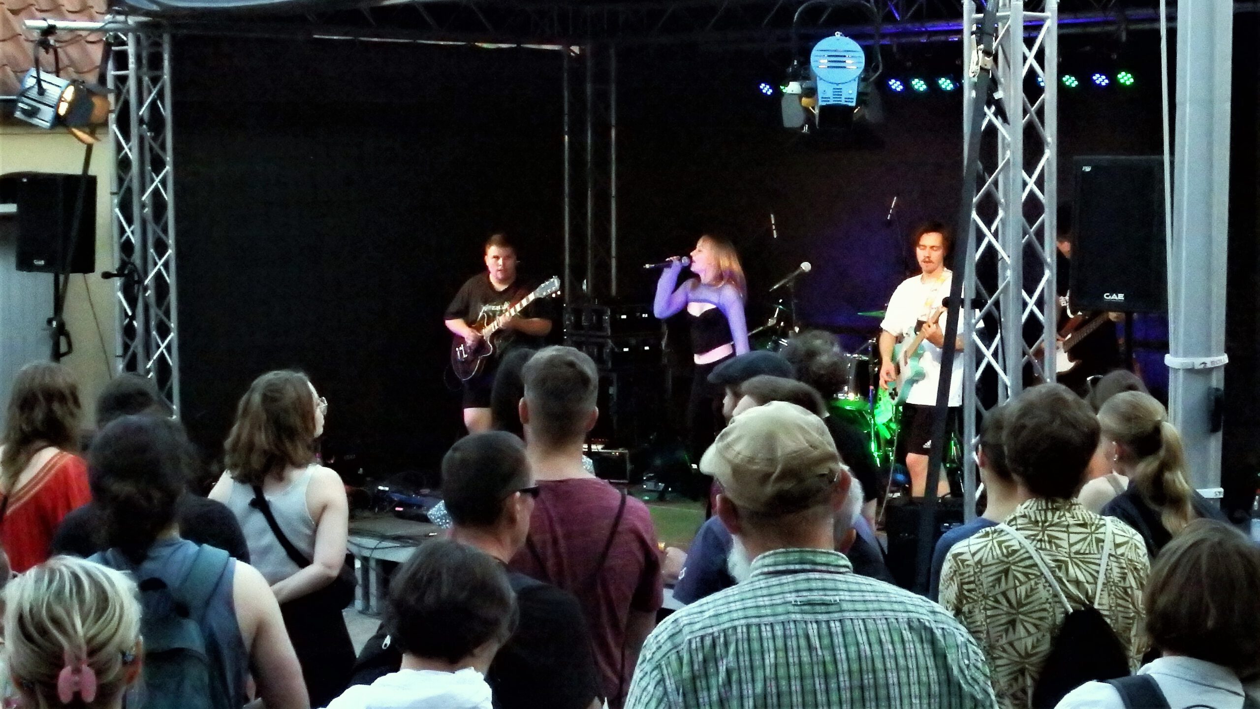 Foto einer Bühne aus dem Publikum heraus, im Vordergrund Zuschauende. Dahinter sind drei Bandmitglieder zu sehen, die äußeren spielen Bass oder E-Gitarre, die Sängerin in der Mitte springt leicht in die Luft.